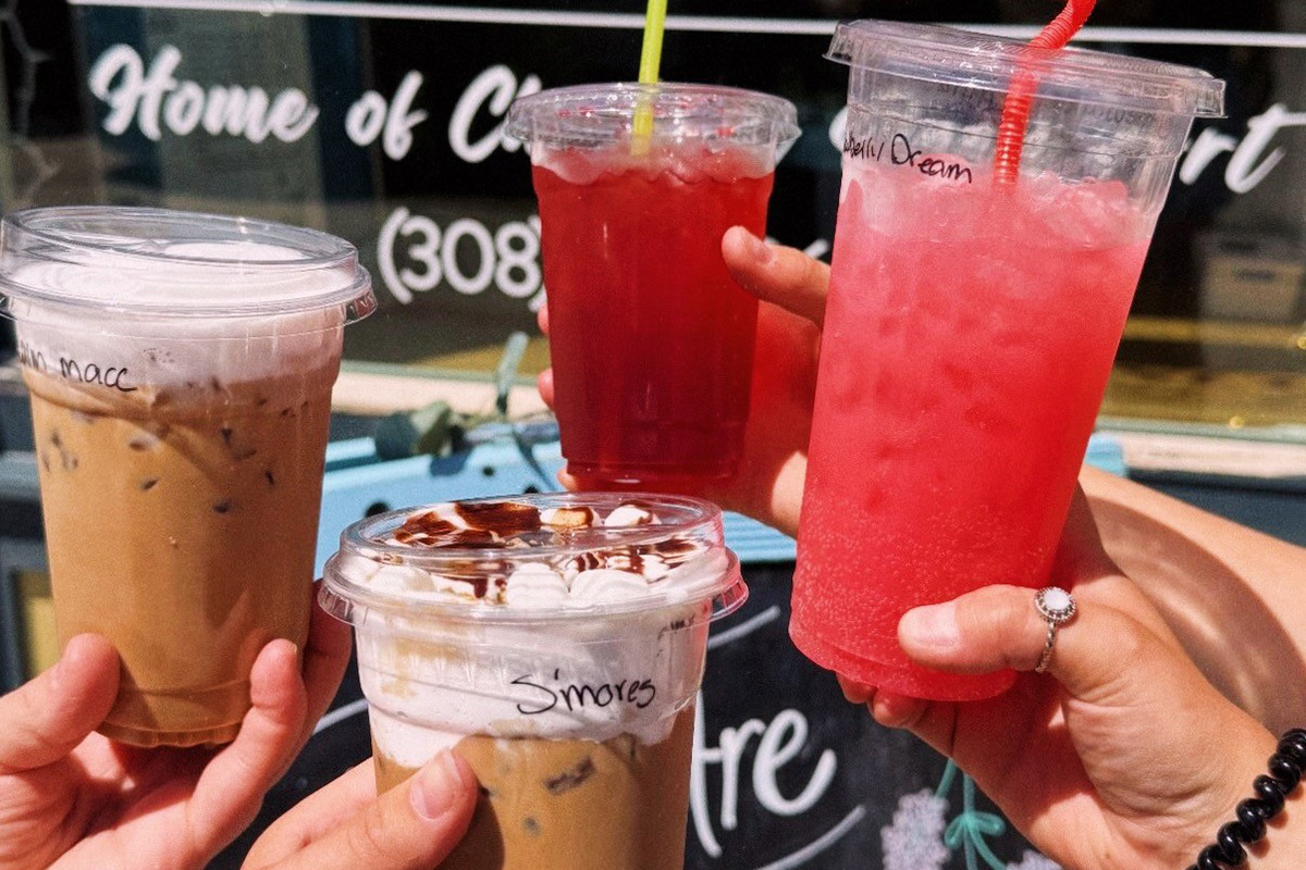 Four hands hold up colorful iced drinks, including coffee and bright red and pink beverages, in front of a café window.