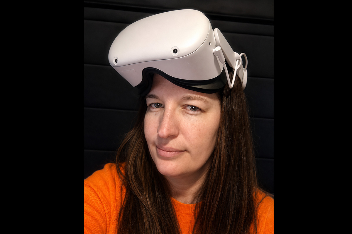 A person with long brown hair wearing a white virtual reality headset pushed up onto their forehead looks at the camera with a slight smile. They are wearing a bright orange sweater and are seated against a dark, padded wall background.
