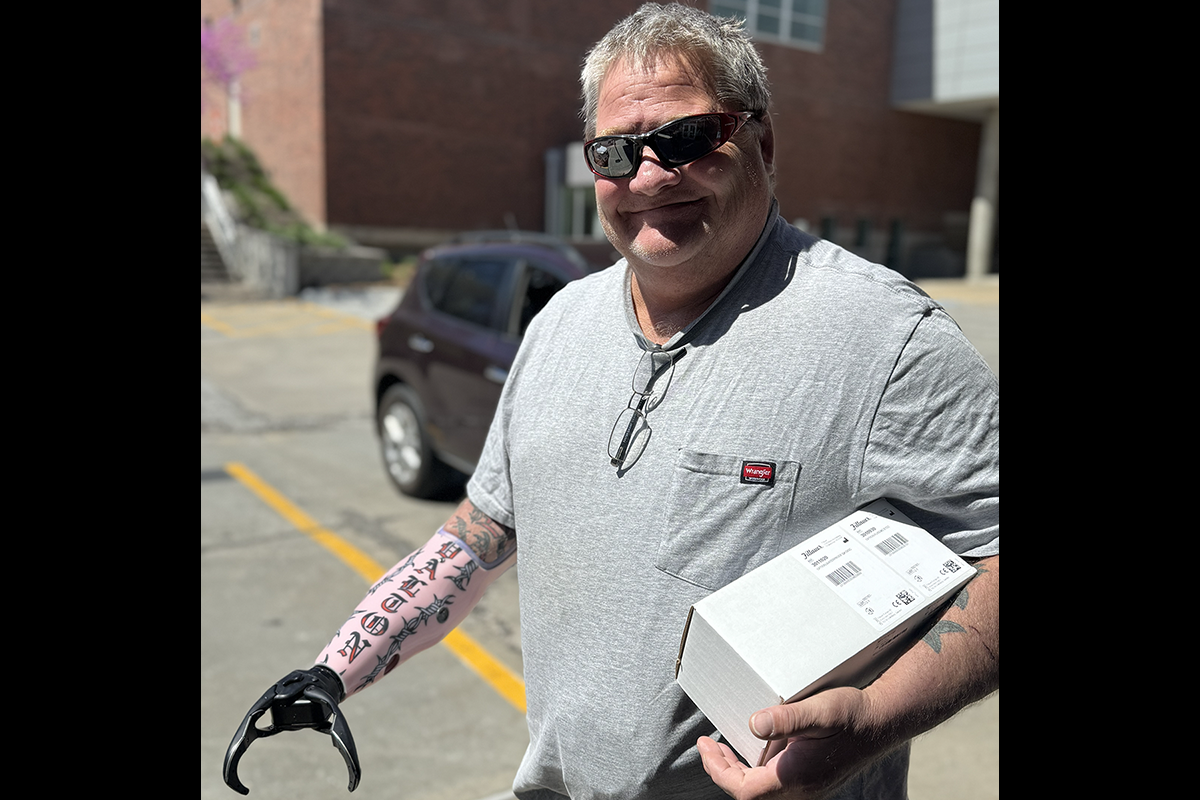 A smiling man wearing sunglasses holds a box while standing outdoors. He has an adaptive prosthetic arm with a hook attachment.
