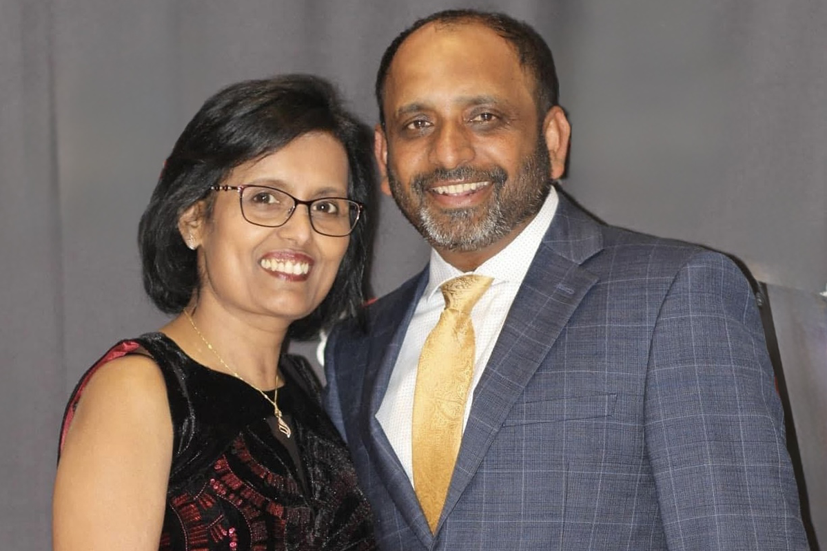A smiling couple poses together at a formal event, with the woman wearing a black and red dress and the man wearing a blue suit with a gold tie.