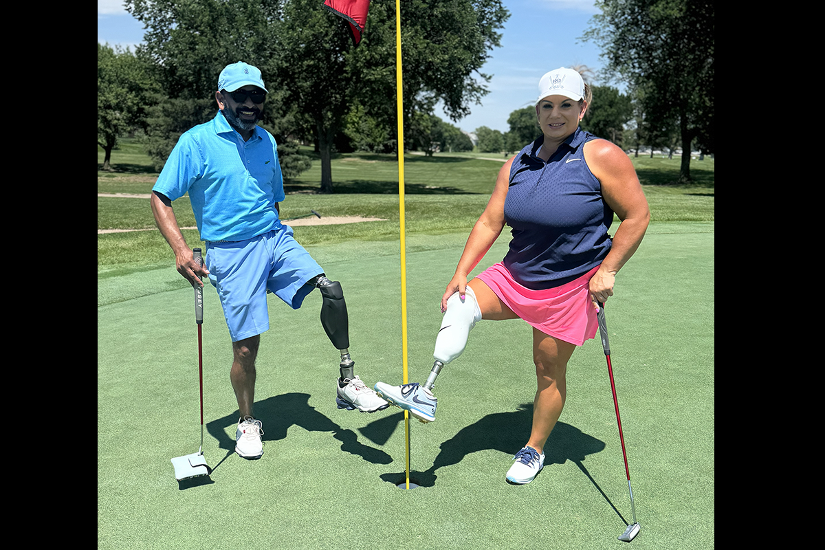 A man and woman with prosthetic legs pose with golf clubs beside the flagstick on a putting green.