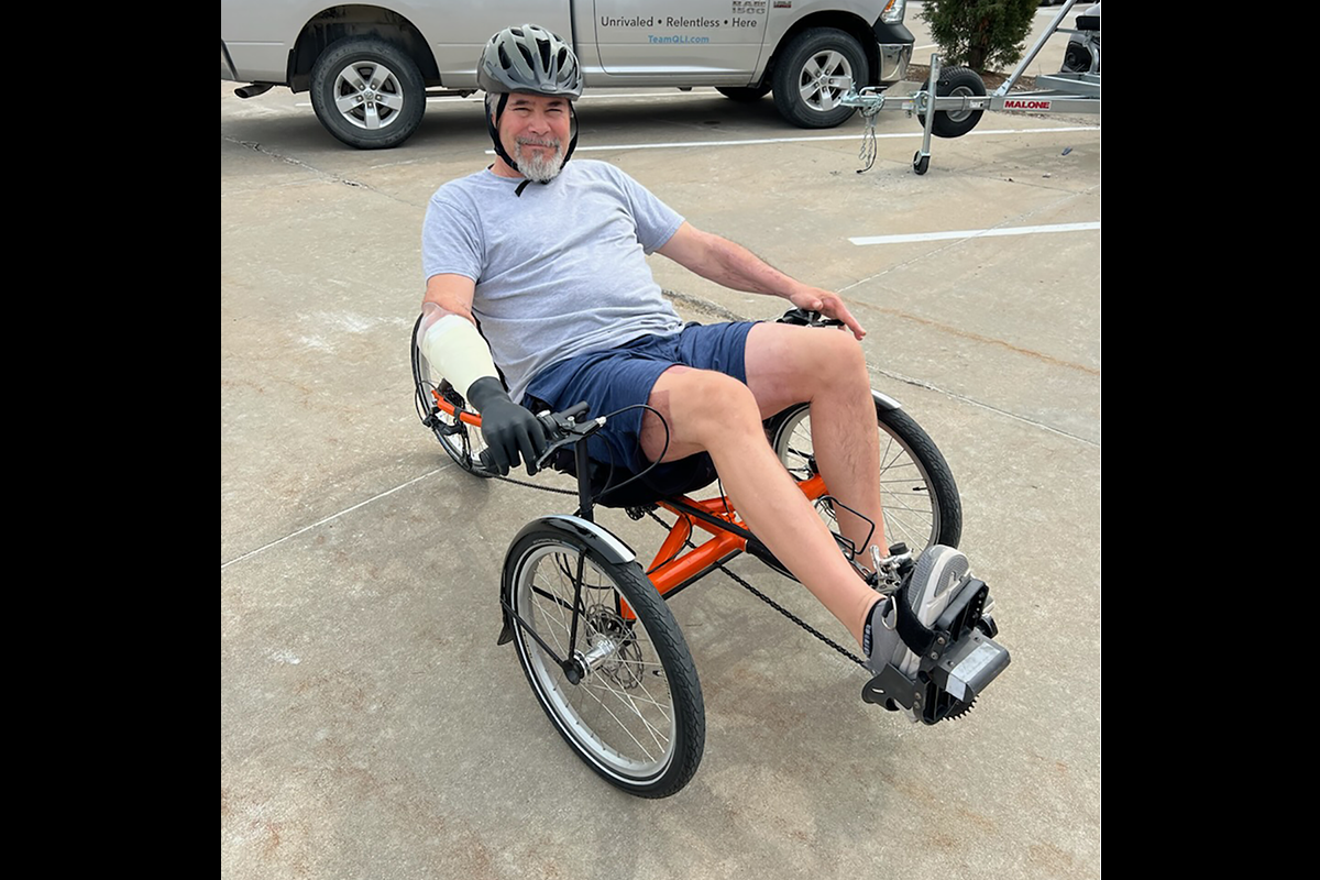 A smiling man wearing a bicycle helmet rides a recumbent tricycle with adaptive hand controls in a parking lot.