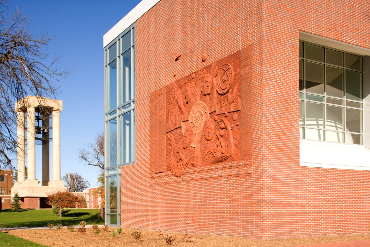 Exterior of Bruner Hall at the University of Nebraska at Kearney, featuring a large sculptural relief on the red-brick facade, with the campus bell tower in the background.