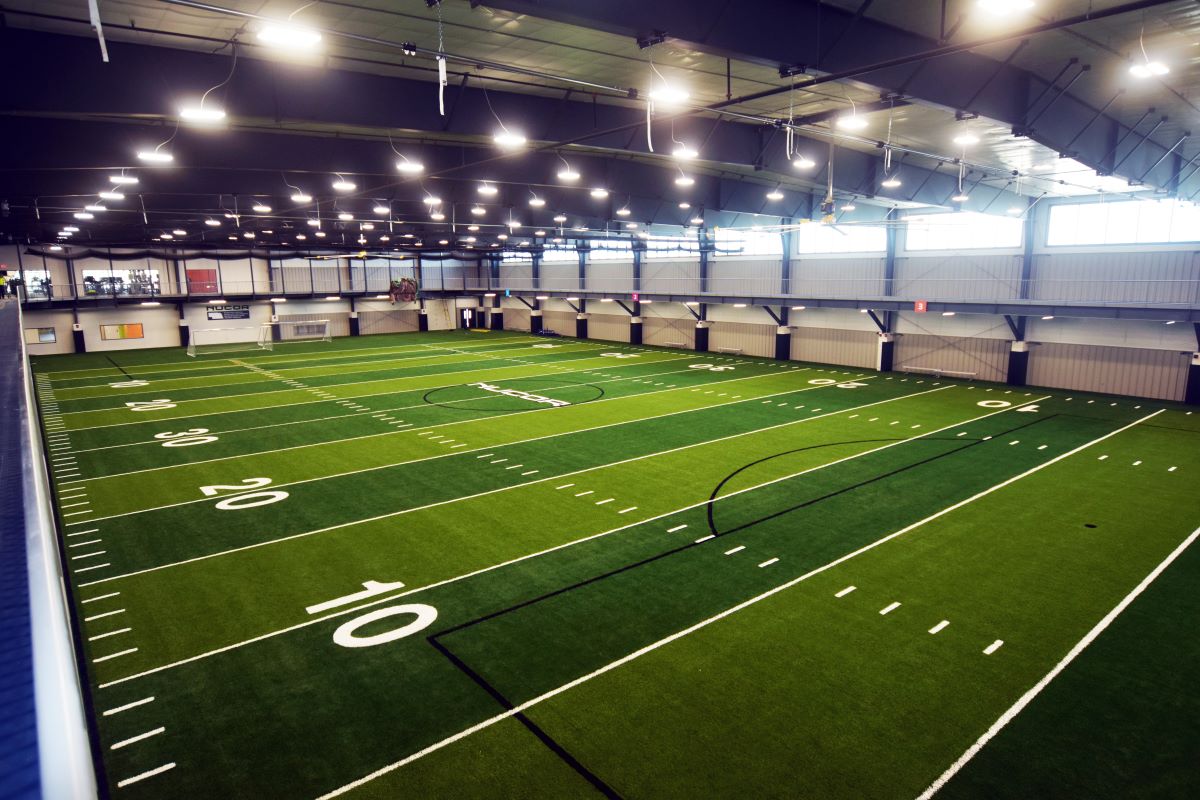 Indoor athletic field house featuring green artificial turf marked with yard lines and field numbers beneath a high ceiling.