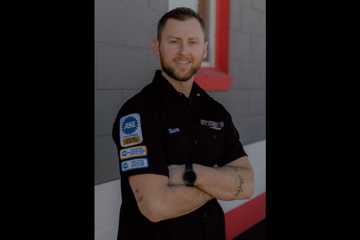 A man wearing a black Eitzen’s Automotive work shirt with certification patches stands with his arms crossed outside a gray building with red trim.