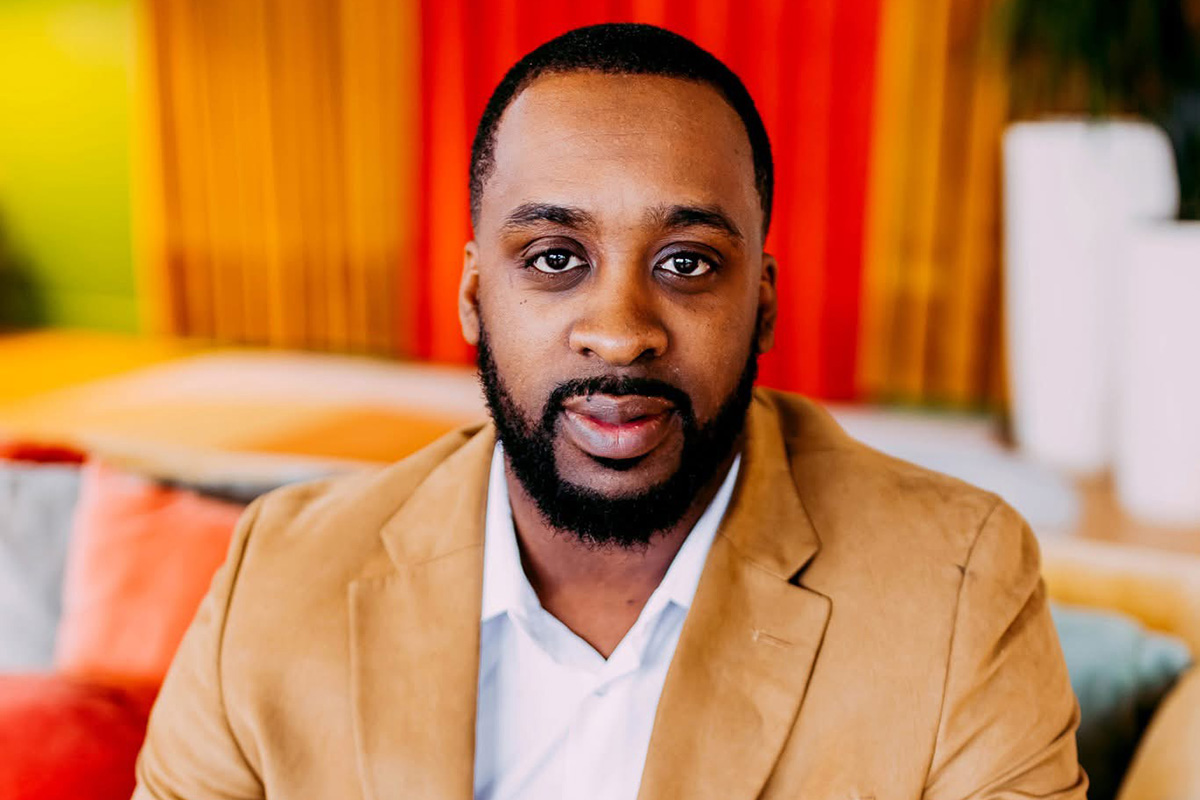 A man with short black hair and a neatly trimmed beard looks directly at the camera, wearing a tan blazer over a white collared shirt. Colorful orange, yellow and red accents and upholstered seating are visible in the softly blurred background.