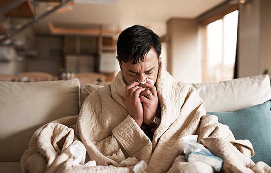 a man sits on his couch, wrapped in a blanket and wiping his nose with a tissue
