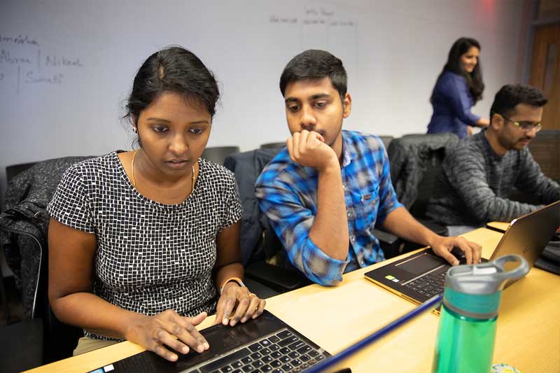 Three students gather around a laptop during a MIS Capstone course meeting