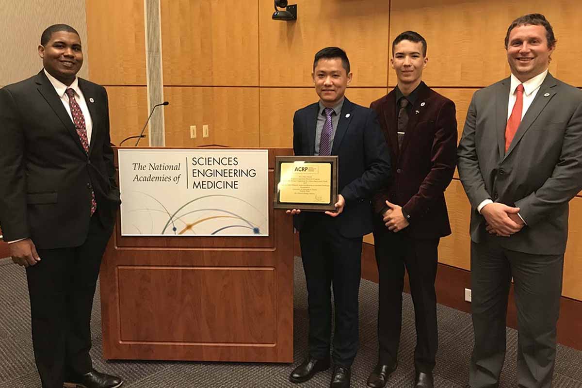 Joseph Harris, Chenyu "Victor" Huang Ph.D., Alexander Nguyen, and Christopher Kelley pose with an award from the Airport Collaborative Research Program (ACRP) next to a podium with a banner that reads "The National Academies of Sciences, Engineering, and Medicine."