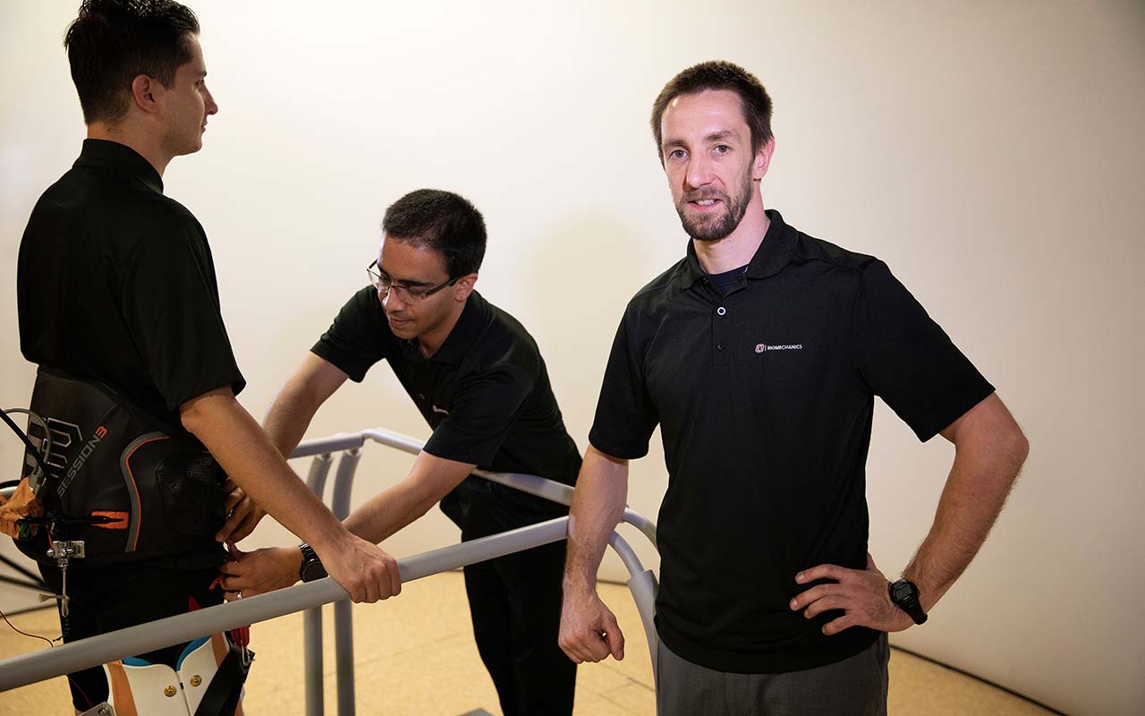 Philippe Malcolm, Ph.D., stands next to two students hooking up an exosuit device that, when used, can reduce the amount of energy required to walk or run.