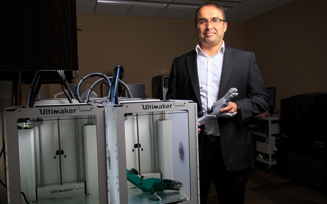 Assistant Professor Jorge Zuniga, Ph.D. holds a plastic prosthetic arm and stands next to a group of 3D printers in UNO's Biomechanics Research Building. Zuniga leads research into the development and production 3D-printed prosthetic limbs for children.