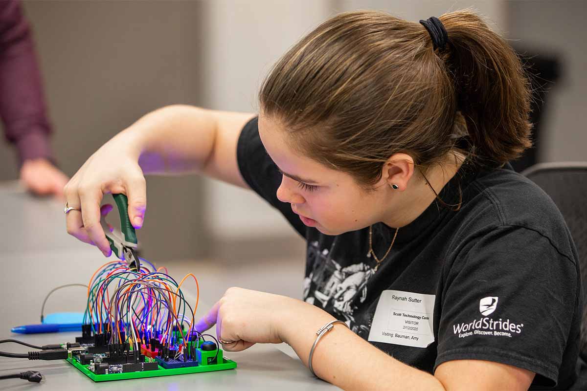 A student holds wire cutters next to a board with wires, lights, and buttons. She carefully takes direction from others to ensure she cuts the right wire in order to defuse a simulated bomb.