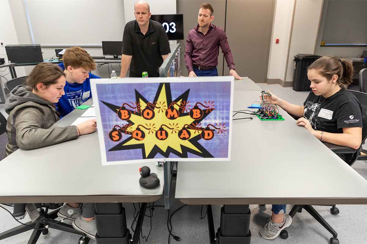 A student holds wire cutters next to a board with wires, lights, and buttons. She carefully takes direction from others to ensure she cuts the right wire in order to defuse a simulated bomb.