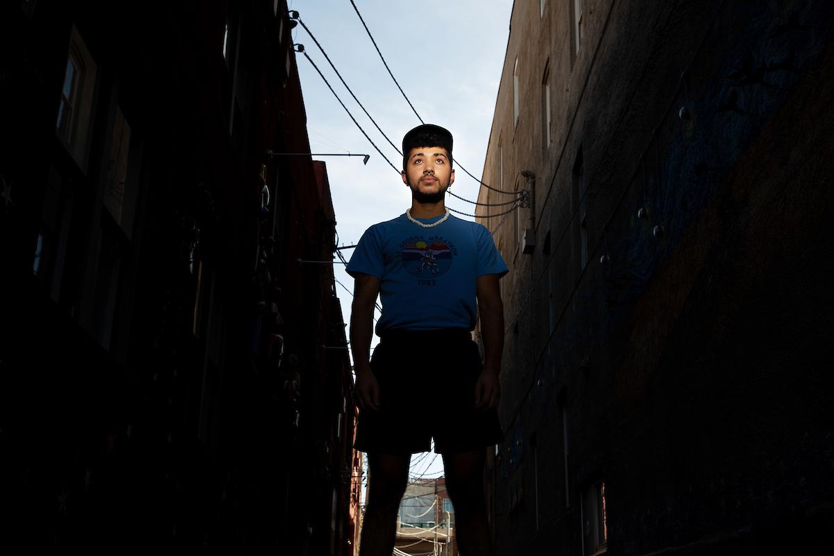 Dakota Chambers poses for a photo in between two dark buildings, his head raised to a clear sky.