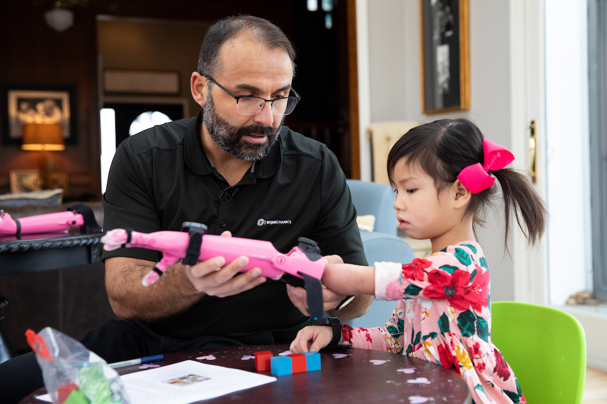 A man in a black shirt assists a young girl with a pink bow in her hair in putting on a pink prosthetic hand.