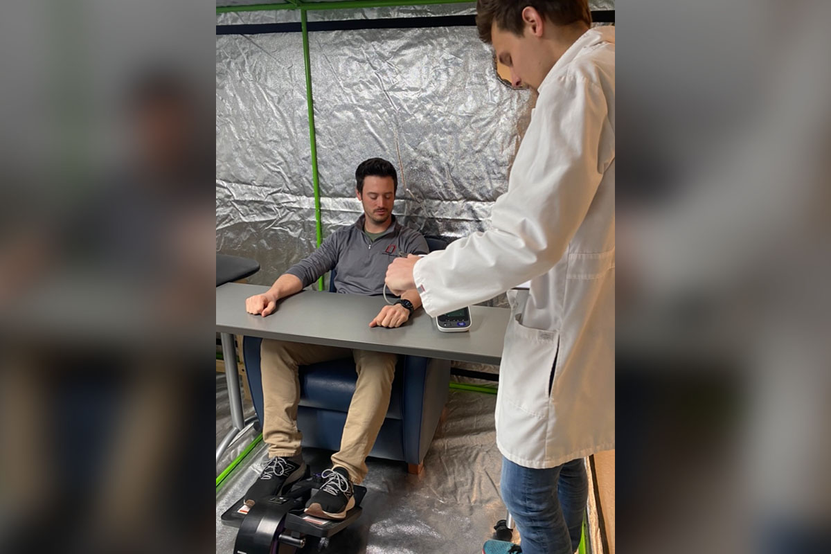 A study participant prepares to pedal on an elliptical while a researcher stands by to take measurements as part of a UNO study into leg movement in environments with relatively higher levels of carbon dioxide, such as an office, classroom, or airline cabin.