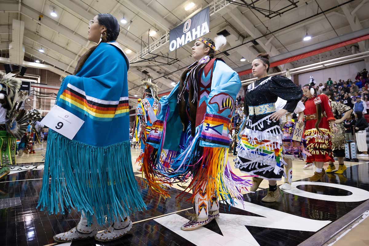 A group of Native American dancers in regalia are shown walking in procession at the Wambli Sapa Memorial Powwow. The dancers are dressed in colorful outfits with fringed garments, beads, and feathers, representing their cultural heritage. The event takes place inside a gymnasium, with spectators visible in the background, highlighting the significance of the gathering.