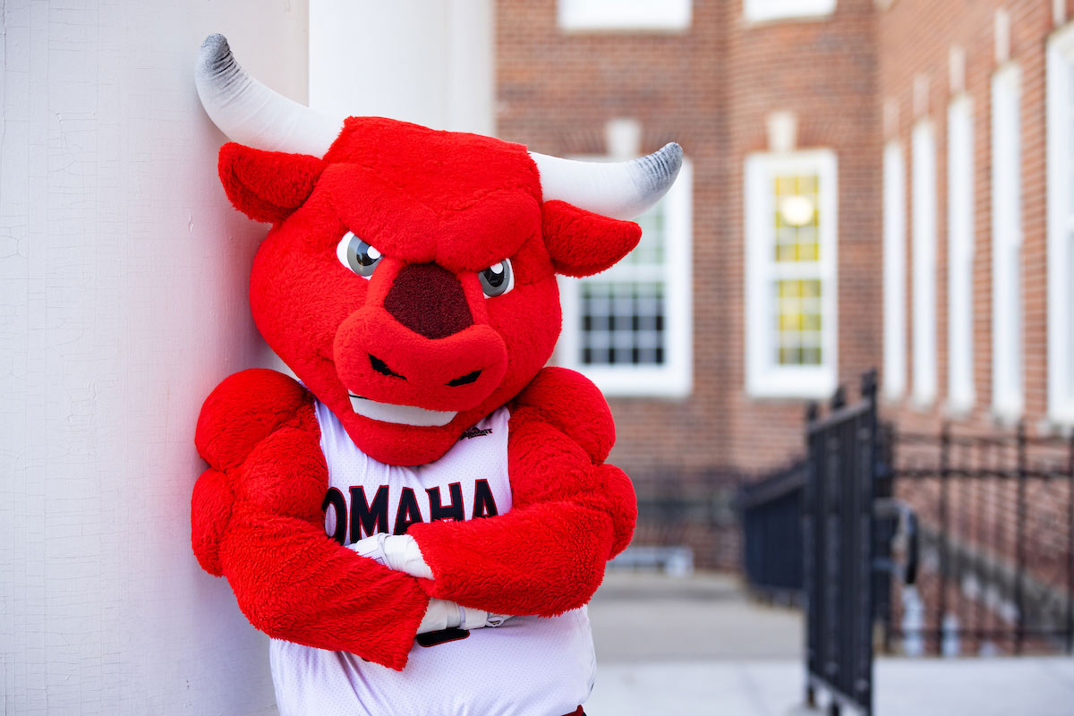 Durango, the red bull mascot for UNO, striking a fierce pose with my arms crossed and a determined look, all while rocking my Omaha jersey in front of a brick building.