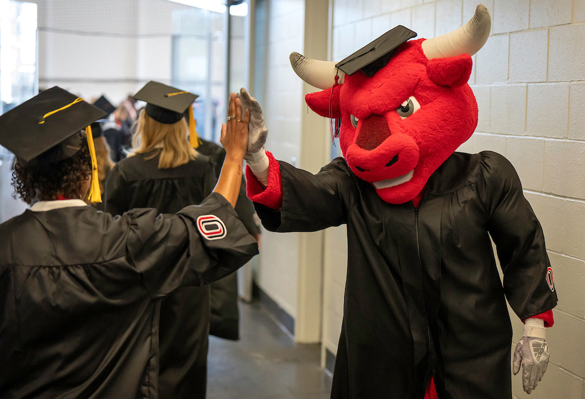 Durango, wearing a graduation cap and gown, high fives a graduating student in cap and gown during the UNO commencement ceremony.