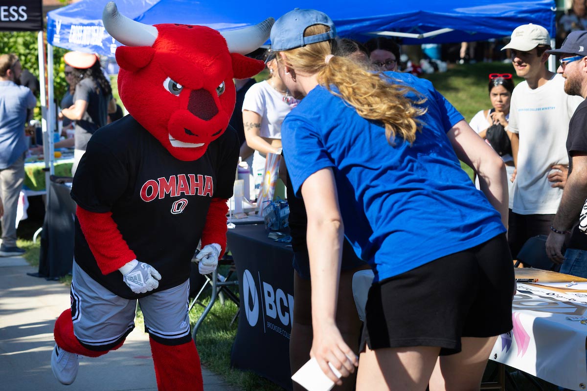 Durango, the red bull mascot, runs toward a student in a blue shirt at MavFest, ready to give a high-five. The scene is filled with other students and event booths in the background.