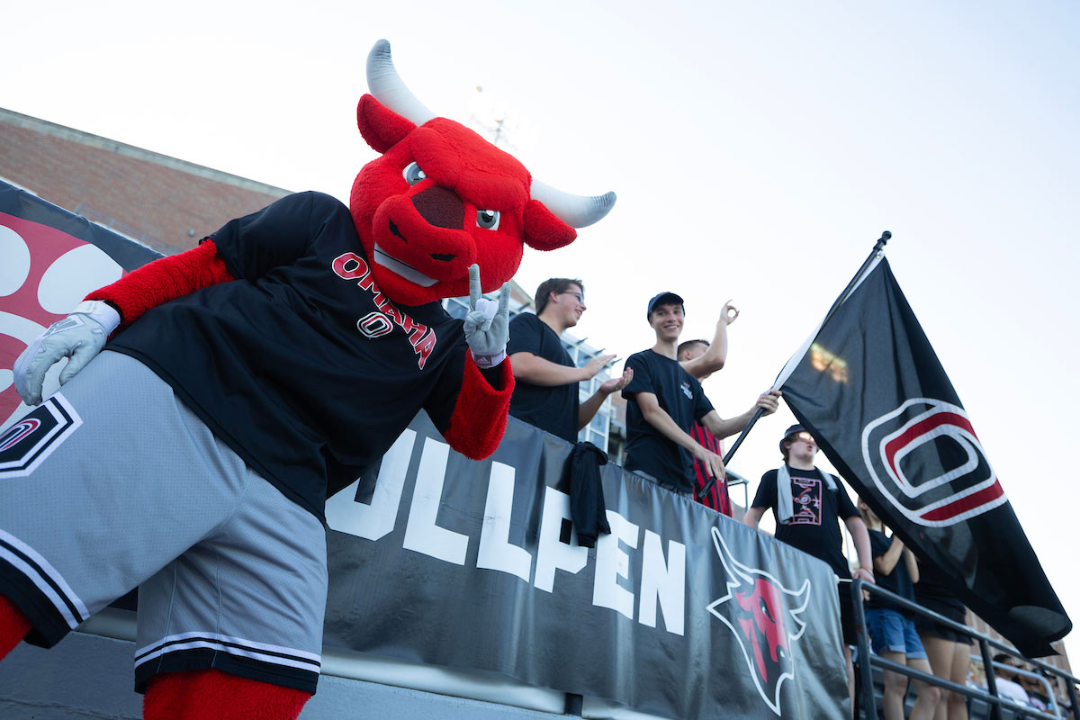 Durango, the UNO mascot, gives a peace sign while standing in front of the fan section during a game. He's wearing a black UNO shirt and gray shorts, surrounded by cheering fans holding UNO flags.