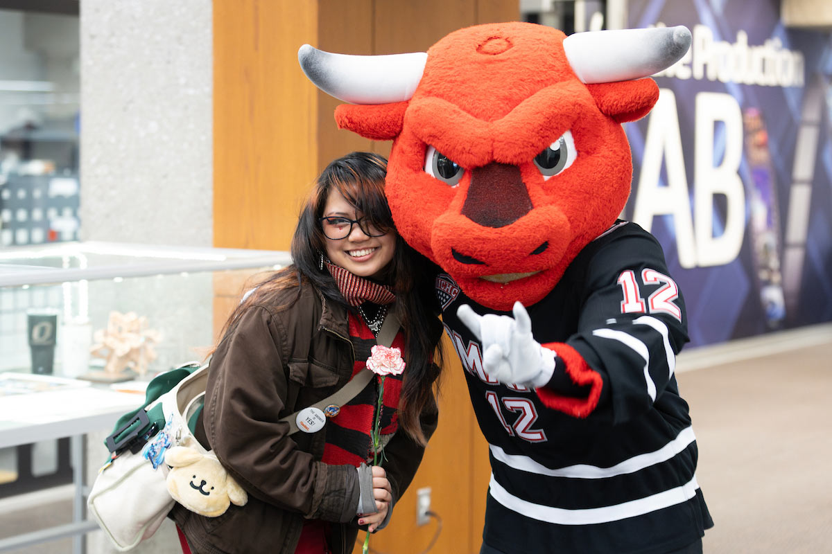 Durango, the red bull mascot, poses for a photo with a smiling student holding a flower on Valentine’s Day. Durango is wearing a UNO hockey jersey, giving a peace sign while the student stands beside him, wearing a red striped scarf.