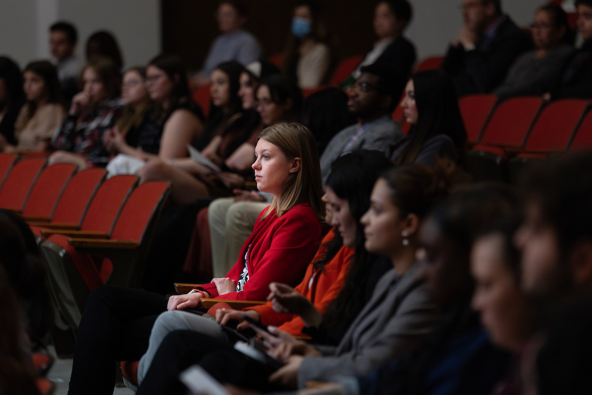 A focused audience watches the 2025 Student Honors Convocation at the University of Nebraska at Omaha. A woman in a red blazer is prominently seated in the foreground, with other attendees in the background.