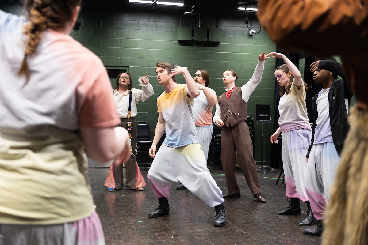 A group of actors in rehearsal clothing practices expressive movements onstage during a Alice in Wonderland run-through, with one actor mid-pose at center.