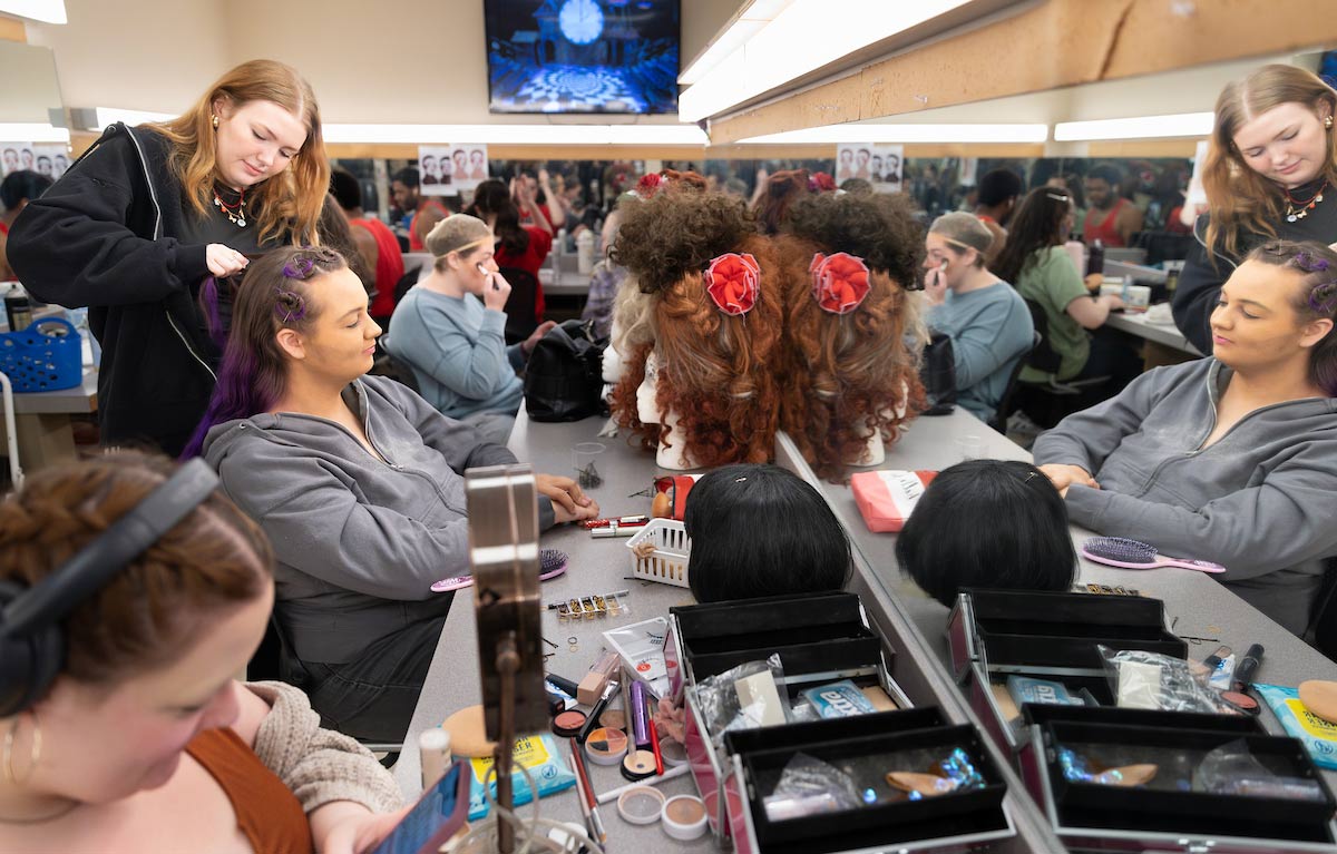 Backstage in the makeup room, cast and crew members prep for the show, styling wigs, applying makeup, and getting ready in front of mirrors filled with costume tools.