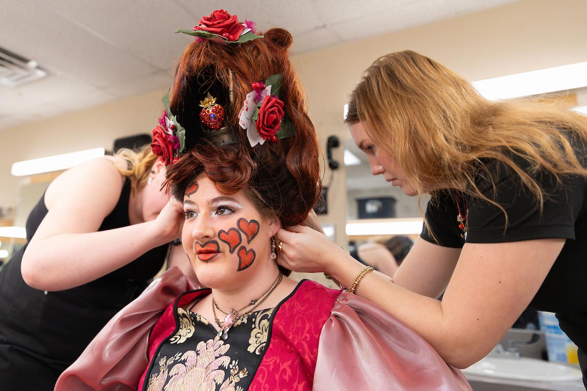 Two crew members help an actor transform into the Queen of Hearts, adjusting an elaborate wig adorned with roses and cards, while the actor smiles in full costume and makeup.
