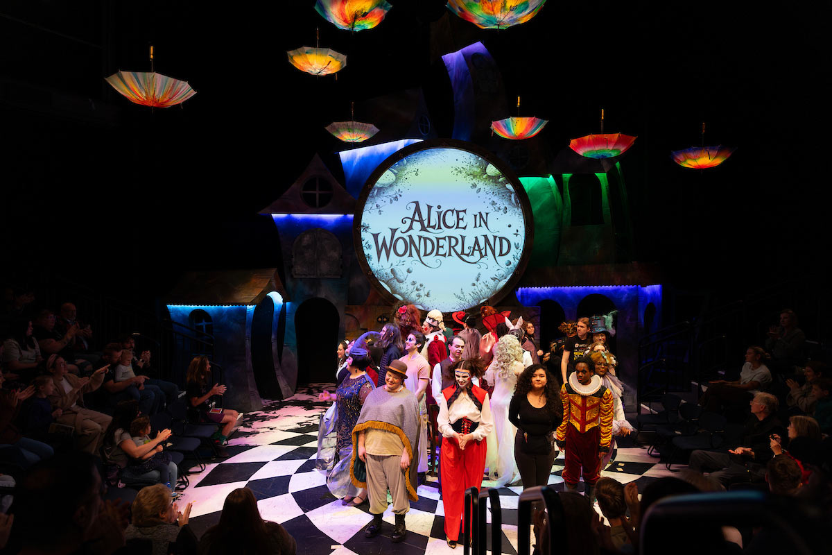 The full cast of Alice in Wonderland stands on stage for curtain call, surrounded by colorful lighting, whimsical set pieces, and a cheering audience.