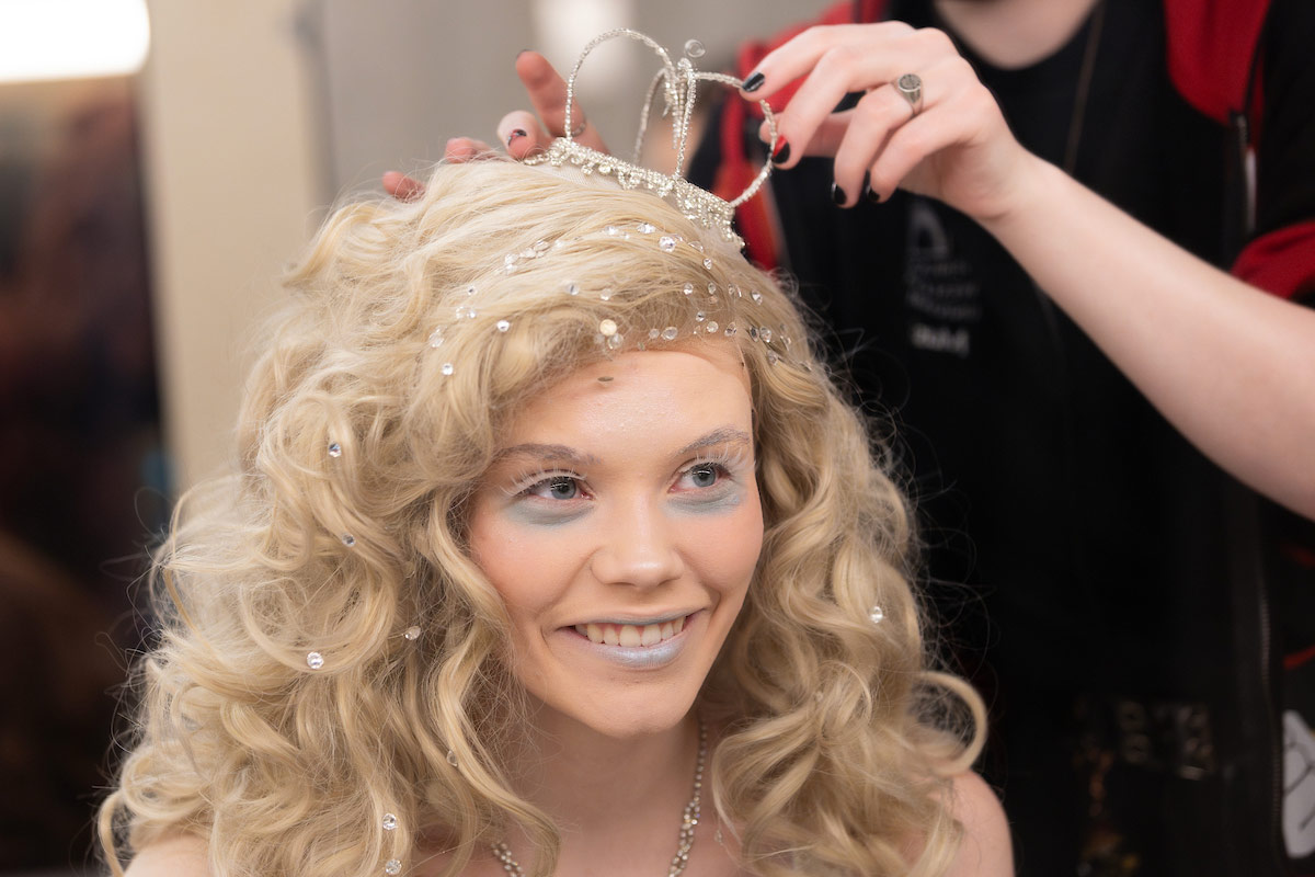 An actor portraying the White Queen smiles while a crew member places a delicate crown on her sparkly blonde wig during pre-show hair and makeup prep.