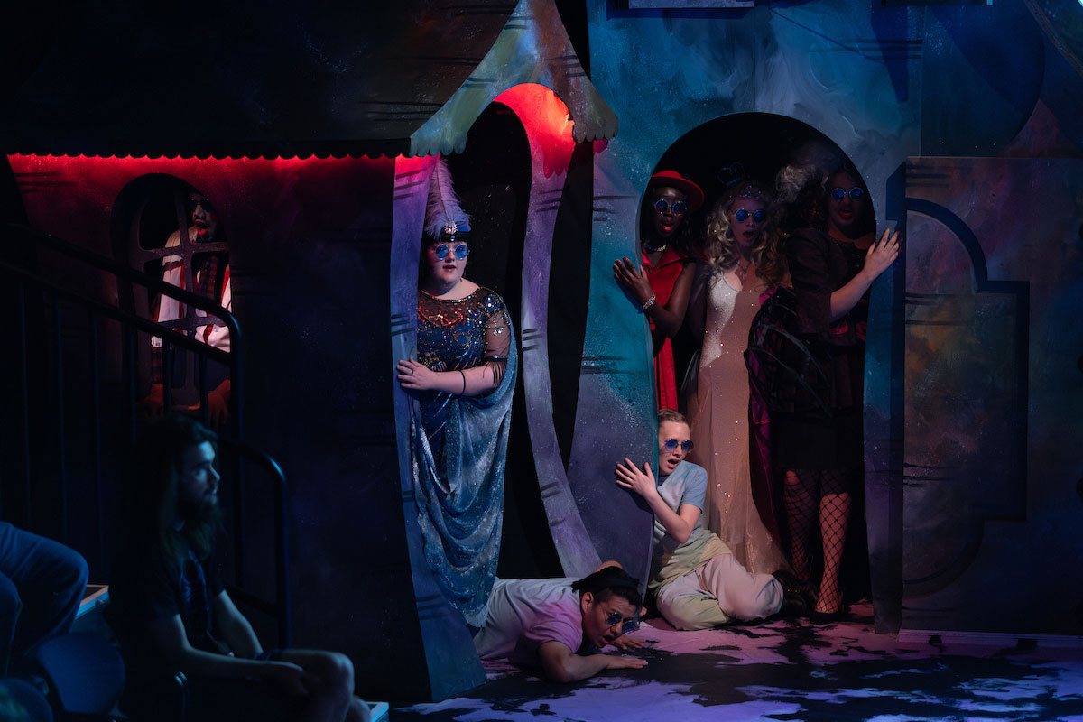 A group of costumed actors in sunglasses peek dramatically out from colorful stage doorways, with one crawling across the floor, under vivid lighting.