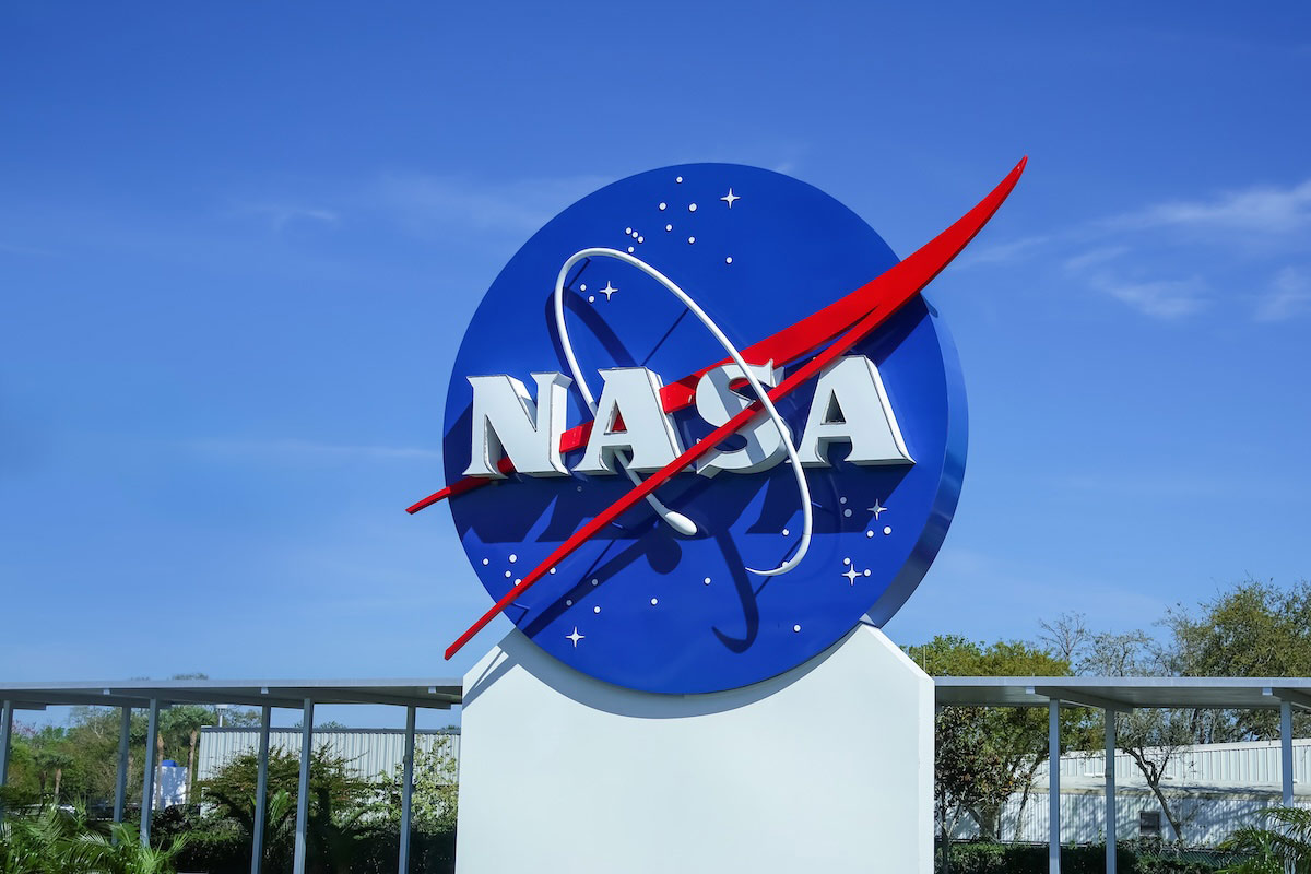 The image shows the NASA logo displayed on a sign outside a facility, with a clear blue sky in the background. The logo consists of a blue circle with white stars, featuring the bold red "NASA" letters crossed by a red diagonal line representing a stylized orbit.