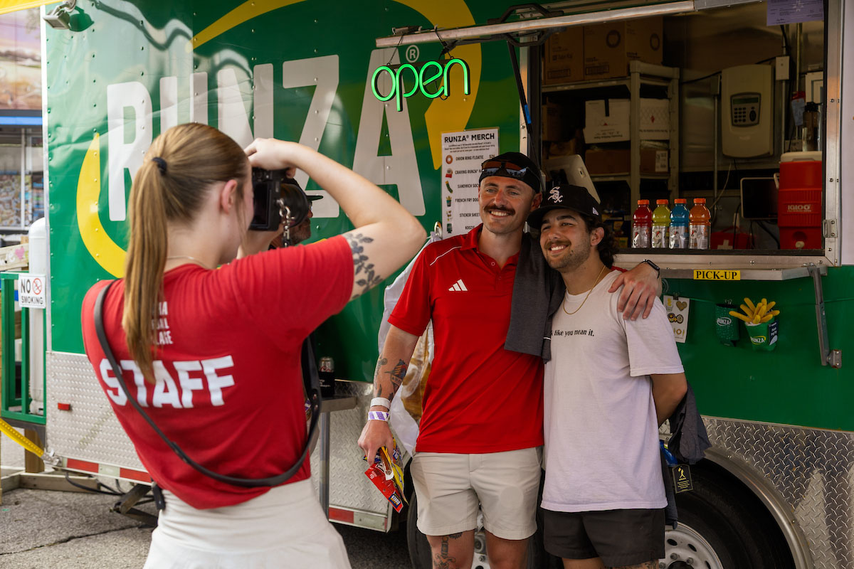 Bella McAtee photographs two smiling men posing in front of a Runza food truck in the Men's College World Series Baseball Village.