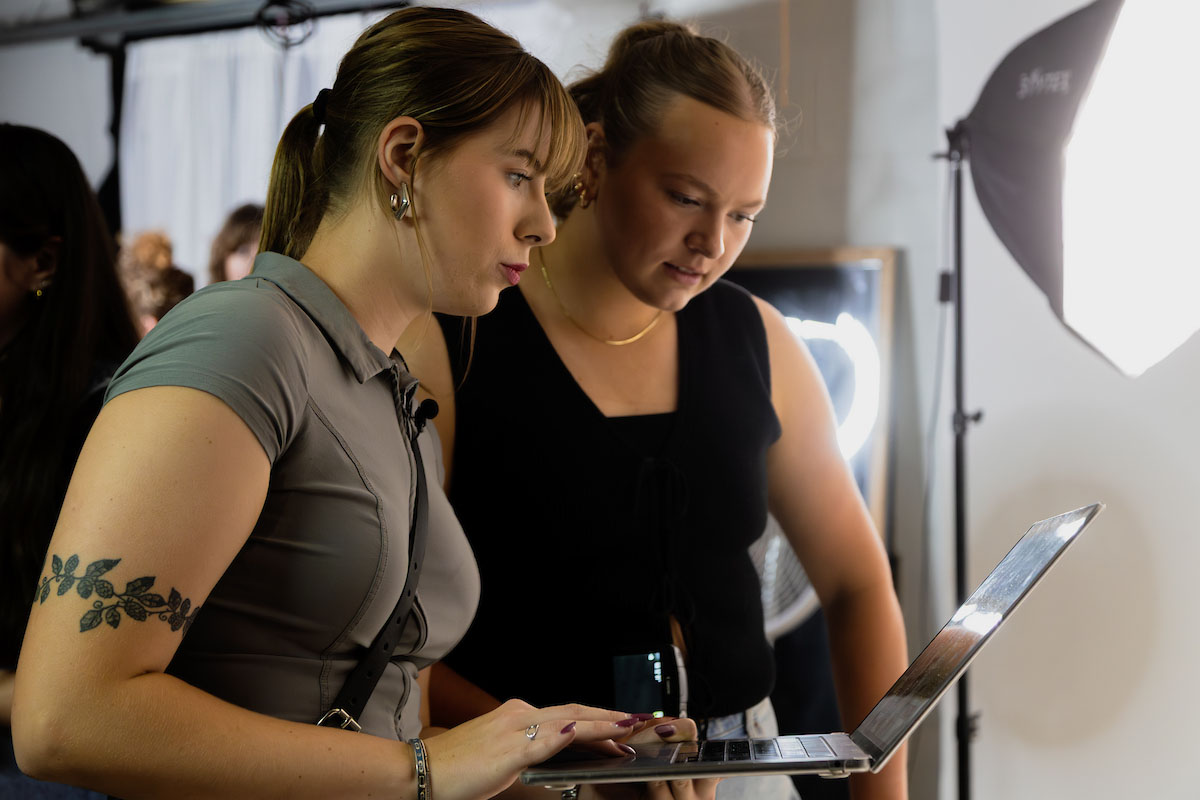 Bella McAtee and a colleague review photos on a laptop during a fashion photo shoot, with studio lights in the background.