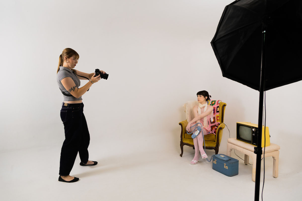 Bella McAtee photographs a seated model in a vintage-styled photo studio with props including a rotary phone and old TV.