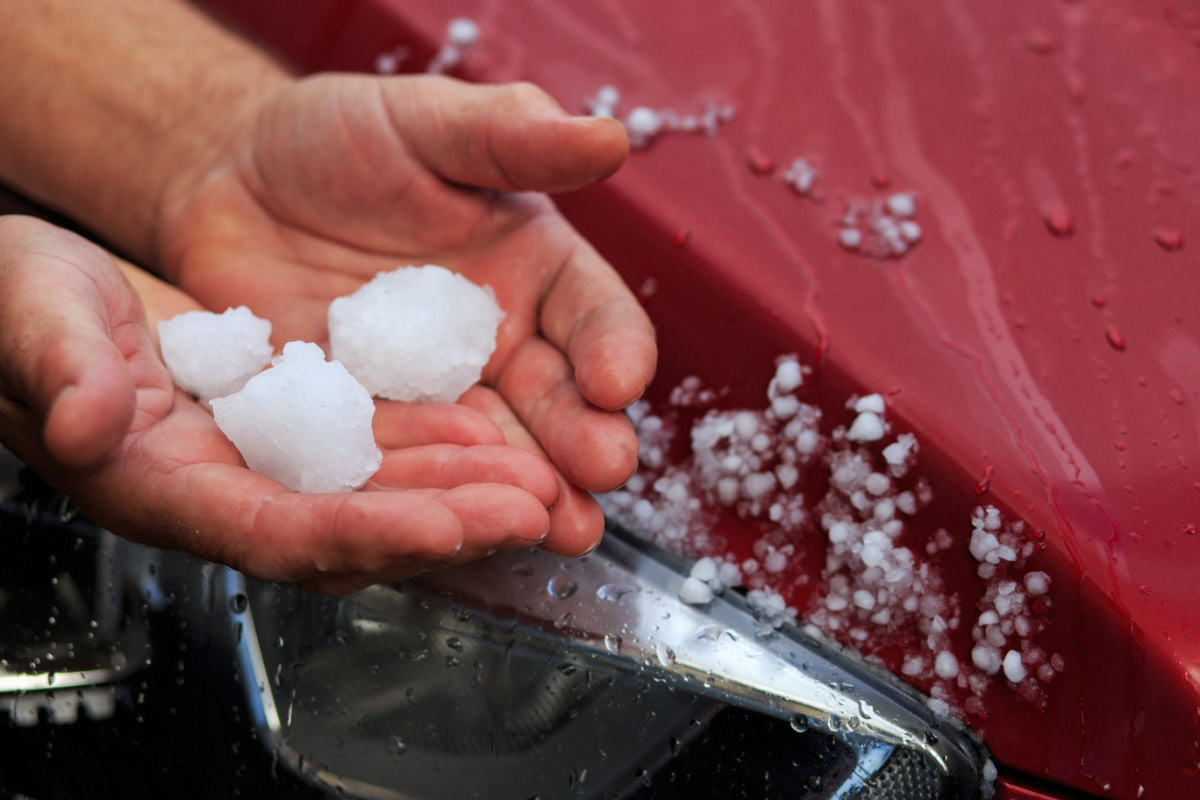 A person holds several large hailstones in their hands next to a red car, where smaller hailstones are scattered across the wet surface.