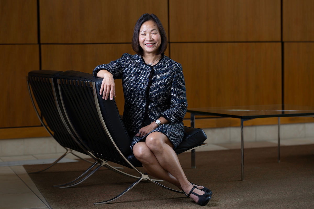 Chancellor Joanne Li sits on a modern black chair, smiling confidently in a professional dark suit, with a wood-paneled wall in the background. 