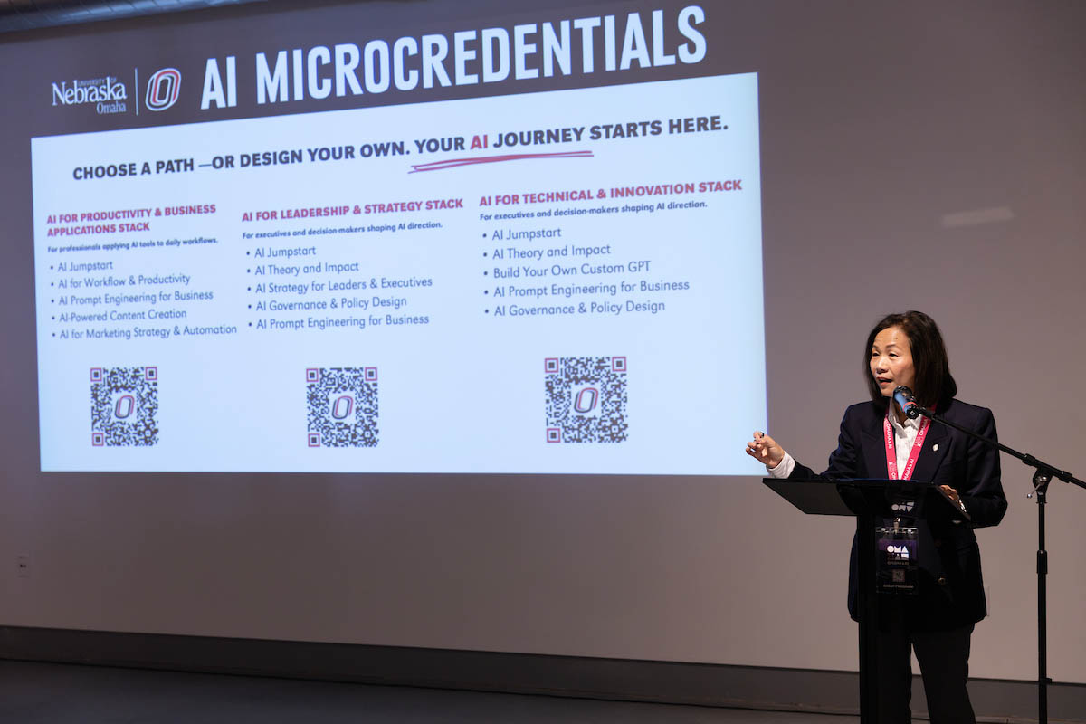 Chancellor Li stands at a podium presenting in front of a projected slide titled "AI Microcredentials" listing course options.
