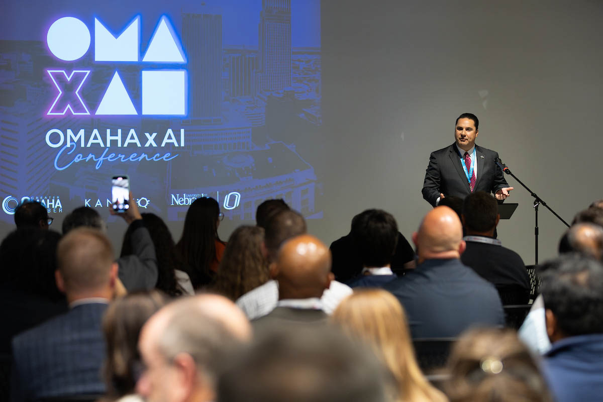 Heath Mello speaks at a podium in front of a projected "OMA x AI Conference" screen, facing a seated audience.