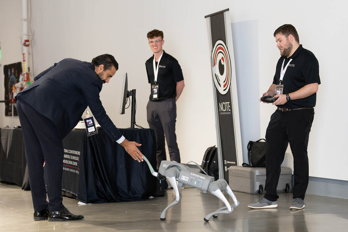 A man in a suit leans down to touch a robotic dog, while two men in branded black shirts observe; one holds a remote control.