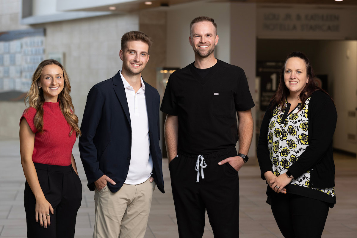 Alyssa Cave, TJ Andreasen, Dr. Landon Peterson, and Lainey Kula stand together inside Mammel Hall at the University of Nebraska at Omaha, smiling for a group photo. The three UNO MBA alumni collaborated with Dr. Peterson on a Capstone project that helped inform the expansion of his orthodontics practice.