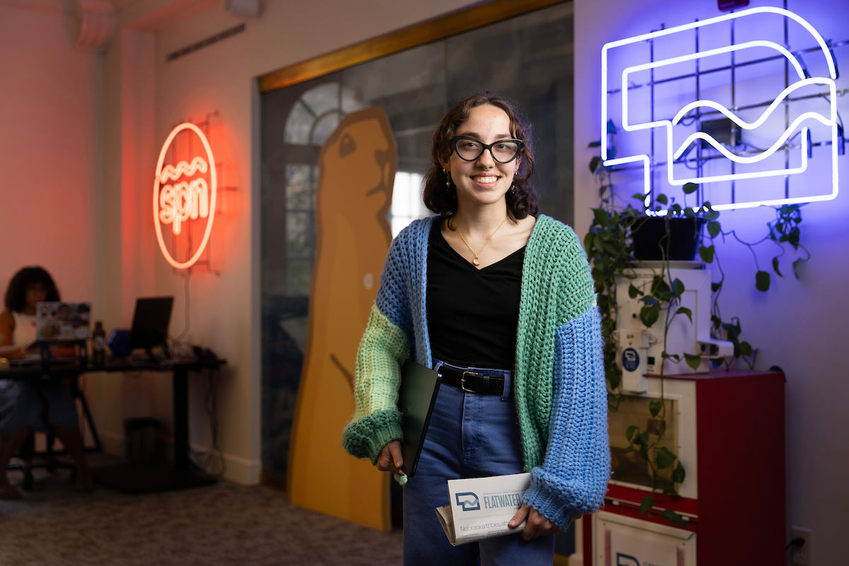 Olivia McArthur, a third-year journalism and media communications major at the University of Nebraska at Omaha, smiles while standing in the Flatwater Free Press office in Omaha. She’s holding her laptop and a Flatwater Free Press folder, wearing glasses, a black shirt, blue jeans, and a blue-and-green knit cardigan. Behind her, colorful neon signs, hanging plants, and a large wall mural create a vibrant newsroom backdrop.