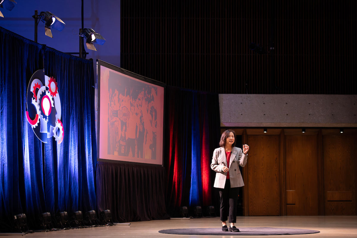 Chancellor Joanne Li speaks on stage during the 2025 State of the University Address, standing in front of a projection screen and a blue curtain featuring a lightbulb and gears graphic. 