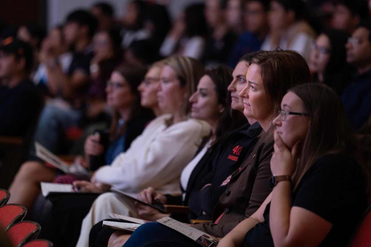 A group of attendees sits closely together, focused on the speaker during the 2025 State of the University Address at UNO. Many hold programs and appear engaged in the presentation. 