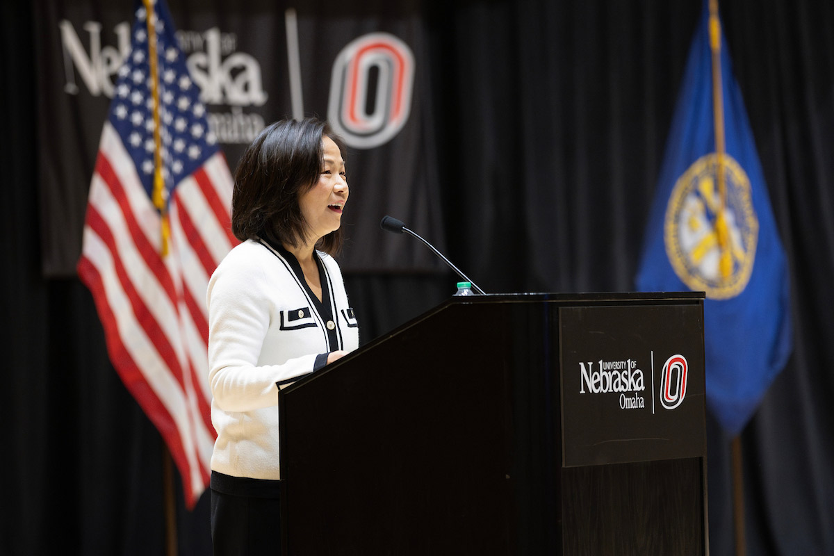 Chancellor Joanne Li speaks at a podium bearing the University of Nebraska at Omaha logo during the Chuck Hagel Symposium in Public Service, with U.S. and Nebraska state flags in the background. 