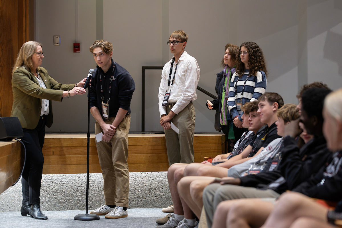 A group of high school students stand in line to ask questions at a microphone during the Chuck Hagel Symposium in Public Service at UNO, as an event facilitator holds the mic. 
