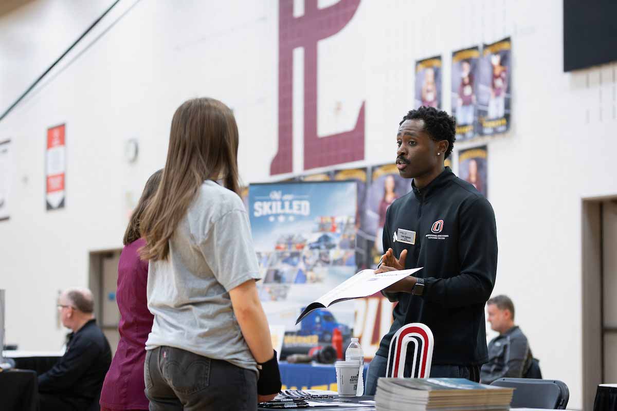 Fabio Gbetanou, a recruiter for the University of Nebraska at Omaha, speaks with a prospective student at a high school career fair. He holds an informational packet while standing behind a UNO display table covered with brochures and a large UNO logo. 