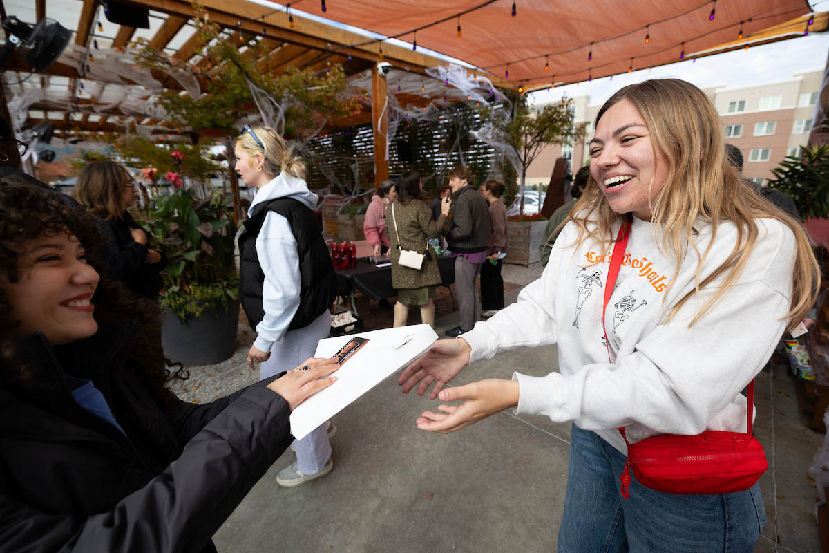 Two students interact at a bustling outdoor market. One wearing a light-colored sweatshirt, smiles as they receives a pizza box from another student.